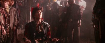 Movie still from “Hook” (1991), directed by Steven Spielberg – A man with a red feather mohawk sits in front of a group of people; Medium shot, Low angle