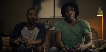 Movie still from “Black Mirror” (2011), created by Charlie Brooker – A couple of men sitting on top of a couch holding remotes; Medium shot, High angle