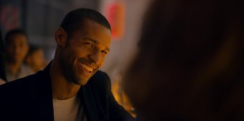 Movie still from “Black Mirror” (2011), created by Charlie Brooker – A man smiling while sitting at a table with other people; Close Up shot, Over the shoulder angle
