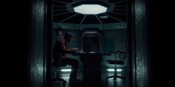 Movie still from “Black Mirror” (2011), created by Charlie Brooker – A man sitting at a table in a room; Wide shot, High angle
