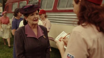 Movie still from “A League of Their Own” (2022), created by Abbi Jacobson – An older woman wearing a purple hat and a purple dress; Medium shot, Over the shoulder angle