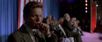 Movie still from “In the Line of Fire” (1993), directed by Wolfgang Petersen – A man sitting in front of an audience while holding his hand to his mouth; Close Up shot, Low angle