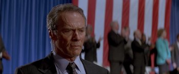 Movie still from “In the Line of Fire” (1993), directed by Wolfgang Petersen – A man in a suit and tie standing in front of an american flag; Close Up shot, Low angle