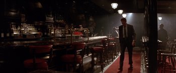 Movie still from “In the Line of Fire” (1993), directed by Wolfgang Petersen – A man standing in front of a bar with red stools; Wide shot, Low angle