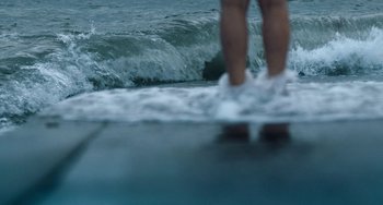 Movie still from “Infamy” (2023), directed by Kuba Czekaj – A person standing on a surfboard in the water; Extreme Wide shot, Low angle