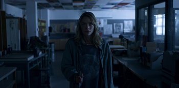 Movie still from “Black Summer” (2019), created by John Hyams – A woman standing in a dark room holding a pen; Medium shot, Low angle