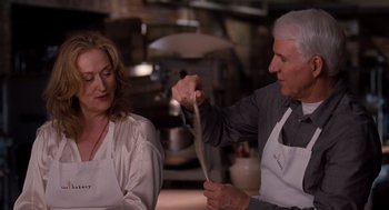 Movie still from “It's Complicated” (2009), directed by Nancy Meyers – A man and a woman in white aprons holding a knife in a kitchen; Medium shot, Over the shoulder angle