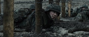 Movie still from “Jarhead” (2005), directed by Sam Mendes – A man in a camouflage suit is crawling under a barbed wire fence; Medium shot, High angle