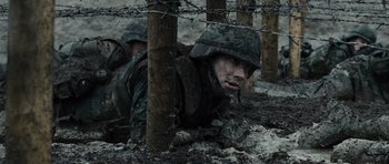Movie still from “Jarhead” (2005), directed by Sam Mendes – A soldier in a helmet is laying in the mud under a barbed wire fence; Medium shot, High angle