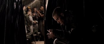 Movie still from “Jarhead” (2005), directed by Sam Mendes – A man sitting on the ground looking at his cell phone; Medium shot, Over the shoulder angle