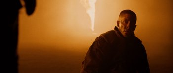 Movie still from “Jarhead” (2005), directed by Sam Mendes – A man standing in front of a wall with a smoke stack in the background; Close Up shot, Over the shoulder angle
