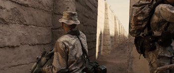 Movie still from “Jarhead” (2005), directed by Sam Mendes – A man with a hat and a camera in front of a brick wall; Medium shot, Over the shoulder angle