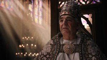 Movie still from “Knightfall” (2017), created by Richard Rayner – An older man wearing a crown in front of a stained glass window; Close Up shot, Low angle