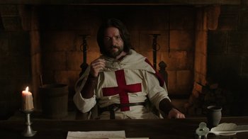 Movie still from “Knightfall” (2017), created by Richard Rayner – A man sitting at a table with a cross on his chest; Medium shot, Low angle