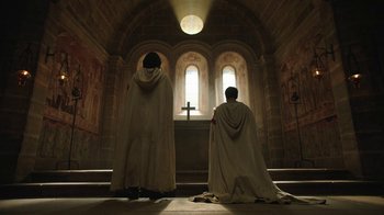 Movie still from “Knightfall” (2017), created by Richard Rayner – Two men in robes kneeling in front of a cross; Wide shot, Low angle