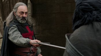Movie still from “Knightfall” (2017), created by Richard Rayner – An old man in a medieval outfit holding a sword; Medium shot, Over the shoulder angle