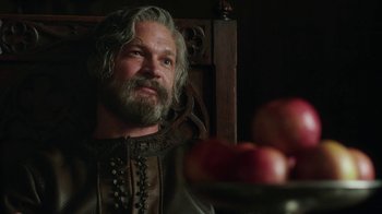 Movie still from “Knightfall” (2017), created by Richard Rayner – A man with long grey hair and a gray beard sitting in front of a bowl of apples; Close Up shot, Over the shoulder angle