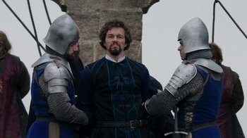 Movie still from “Knightfall” (2017), created by Richard Rayner – Three men in medieval garb standing next to each other in front of a stone wall; Medium shot, Over the shoulder angle