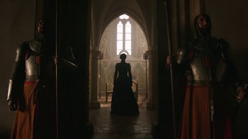 Movie still from “Knightfall” (2017), created by Richard Rayner – A woman dressed in medieval garb stands in front of a window; Wide shot, Low angle