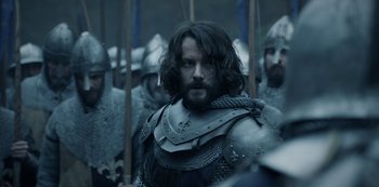 Movie still from “Knightfall” (2017), created by Richard Rayner – A group of men in medieval garb are gathered together; Medium shot, Over the shoulder angle