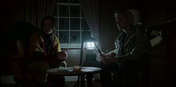 Movie still from “Black Summer” (2019), created by John Hyams – Two people sitting at a table in a dark room; Medium shot, Low angle