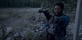 Movie still from “Black Summer” (2019), created by John Hyams – A man holding a gun while standing next to a tree; Medium shot, Over the shoulder angle