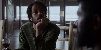 Movie still from “Black Summer” (2019), created by John Hyams – A man sitting in front of a wooden chair with his hand on his chin; Close Up shot, Low angle