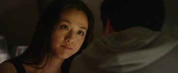 Movie still from “Blackhat” (2015), directed by Michael Mann – A young woman looking at the camera with a man in the background; Close Up shot, Over the shoulder angle