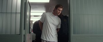 Movie still from “Blackhat” (2015), directed by Michael Mann – A man in a white t - shirt is being escorted by police officers; Medium shot, Low angle