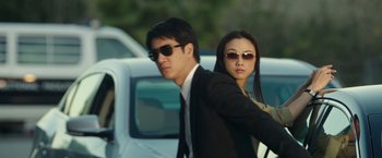Movie still from “Blackhat” (2015), directed by Michael Mann – A man and a woman in suits and sunglasses standing next to each other in front of a white car; Medium shot, Low angle