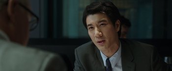 Movie still from “Blackhat” (2015), directed by Michael Mann – A man wearing a suit and tie sitting in front of a window; Close Up shot, Over the shoulder angle