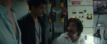 Movie still from “Blackhat” (2015), directed by Michael Mann – A man sitting next to another man on a train; Medium shot, Over the shoulder angle