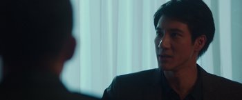 Movie still from “Blackhat” (2015), directed by Michael Mann – A man in a black suit sitting in front of a curtain; Close Up shot, Over the shoulder angle