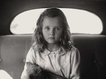 Movie still from “Blonde” (2022), directed by Andrew Dominik – A young girl holding a teddy bear while sitting in the back of a car; Close Up shot, High angle
