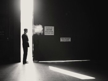Movie still from “Blonde” (2022), directed by Andrew Dominik – A man standing in front of an open door with smoke coming out of it; Wide shot, Low angle
