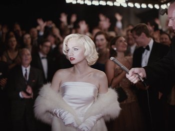 Movie still from “Blonde” (2022), directed by Andrew Dominik – A woman in a white dress holding a microphone in front of a crowd of onlookers; Medium shot, Over the shoulder angle