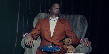 Movie still from “Marcella” (2016), created by Hans Rosenfeldt – A man sitting in a chair holding a teddy bear and a car; Medium shot, Low angle