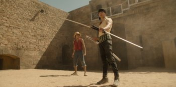 Movie still from “One Piece” (2023), directed by Marc Jobst – A man and a woman are standing in the sand; Wide shot, Low angle