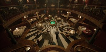 Movie still from “One Piece” (2023), directed by Marc Jobst – A man standing inside of a room with tables and chairs around it; Extreme Wide shot, High angle