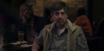 Movie still from “Paddleton” (2019), directed by Alex Lehmann – A man in glasses and a vest sitting at a table with other people; Close Up shot, Over the shoulder angle