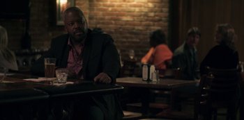 Movie still from “Paddleton” (2019), directed by Alex Lehmann – A man sitting at a table in front of a brick wall; Medium shot, Over the shoulder angle