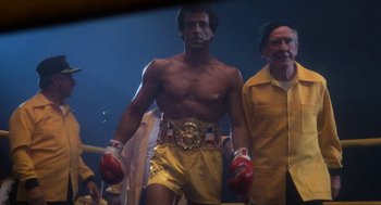 Movie still from “Rocky III” (1982), directed by Sylvester Stallone – A man in yellow trunks and red boxing mitts stands in front of a crowd; Medium shot, Low angle