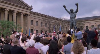Movie still from “Rocky III” (1982), directed by Sylvester Stallone – A crowd of people gathered in front of a statue; Extreme Wide shot, High angle