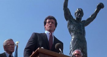 Movie still from “Rocky III” (1982), directed by Sylvester Stallone – A man standing at a podium in front of a statue; Medium shot, Low angle