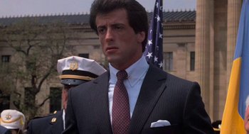Movie still from “Rocky III” (1982), directed by Sylvester Stallone – A man in a suit standing next to an american flag; Close Up shot, Low angle