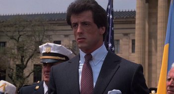 Movie still from “Rocky III” (1982), directed by Sylvester Stallone – A man in a suit standing next to an american flag; Close Up shot, Low angle