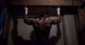 Movie still from “Rocky III” (1982), directed by Sylvester Stallone – A man is doing pull - ups in a gym; Medium shot, Low angle