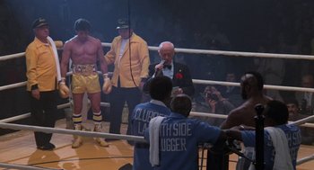 Movie still from “Rocky III” (1982), directed by Sylvester Stallone – A man in a boxing ring with other men; Wide shot, Over the shoulder angle