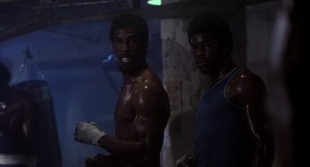 Movie still from “Rocky III” (1982), directed by Sylvester Stallone – Two boxers are posing for a picture in the dark; Medium shot, Low angle