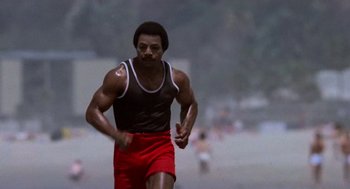 Movie still from “Rocky III” (1982), directed by Sylvester Stallone – A man running on the beach in a black tank top and red shorts; Medium shot, Low angle